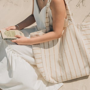 May include: A woman wearing a white dress sits on the sand holding a book and a large striped tote bag.