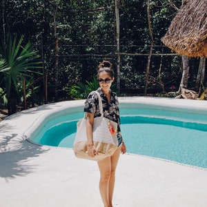 Puede incluir: Una mujer con una camisa de flores blancas y negras y gafas de sol est&aacute; de pie junto a una piscina sosteniendo una bolsa de mano tejida blanca y marr&oacute;n.