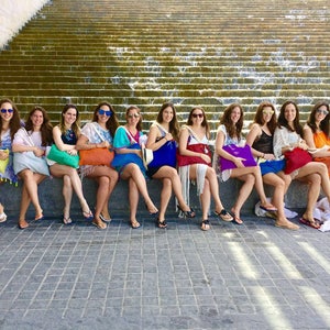 May include: A group of 12 women wearing sunglasses and summer dresses are sitting on a stone ledge in front of a cascading waterfall. They are all holding colourful tote bags.