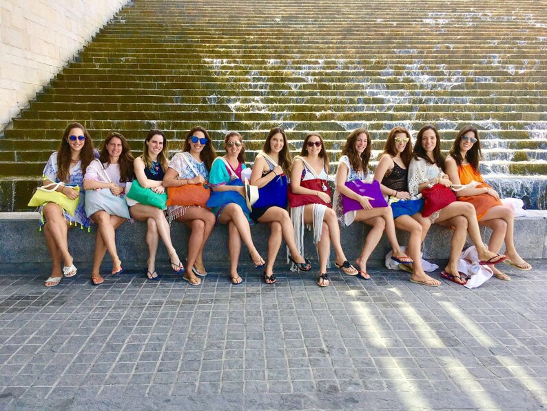 May include: A group of 12 women wearing sunglasses and summer dresses are sitting on a stone ledge in front of a cascading waterfall. They are all holding colourful tote bags.
