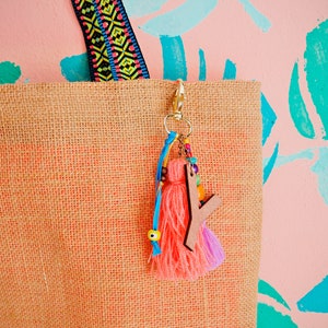 May include: A natural jute tote bag with a pink and blue tassel keychain. The keychain features a wooden letter charm and colorful beads.