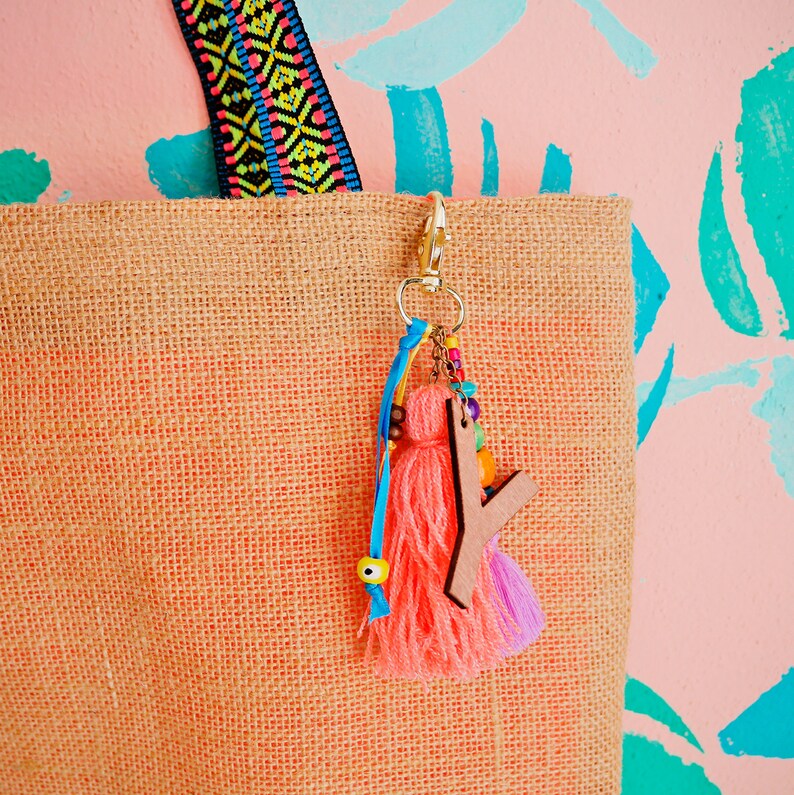May include: A natural jute tote bag with a pink and blue tassel keychain. The keychain features a wooden letter charm and colorful beads.