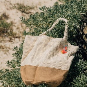 Puede incluir: Una bolsa de mano tejida de color blanco y marr&oacute;n con asas de cuerda y un abalorio de borla de colores. La bolsa est&aacute; sentada sobre un lecho de follaje verde.
