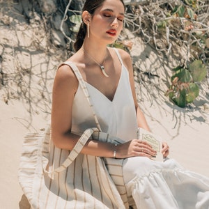May include: A woman wearing a white tank top and white pants sits on a sandy beach with a striped tote bag and a book. She is wearing silver earrings and a necklace with a silver pendant.
