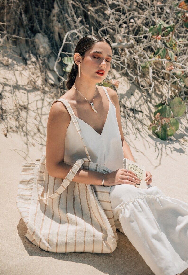 May include: A woman wearing a white tank top and white pants sits on a sandy beach with a striped tote bag and a book. She is wearing silver earrings and a necklace with a silver pendant.