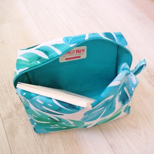 May include: A turquoise and white zippered pouch with a tropical leaf print. The pouch is open and a book is partially inside. The pouch has a label that reads "Paso Bien Happily Made in Cancun".
