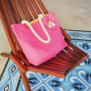 May include: A pink canvas tote bag with a yellow trim and white rope handles. The bag is sitting on a wooden chair with a blue and white tiled floor.