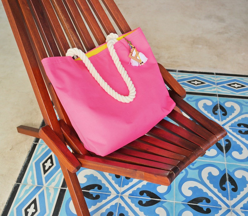 May include: A pink canvas tote bag with a yellow trim and white rope handles. The bag is sitting on a wooden chair with a blue and white tiled floor.