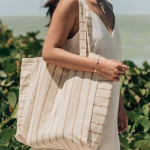 May include: A woman is carrying a large, striped tote bag with tan and white stripes. The bag is made of a natural fabric and has a relaxed, casual style.