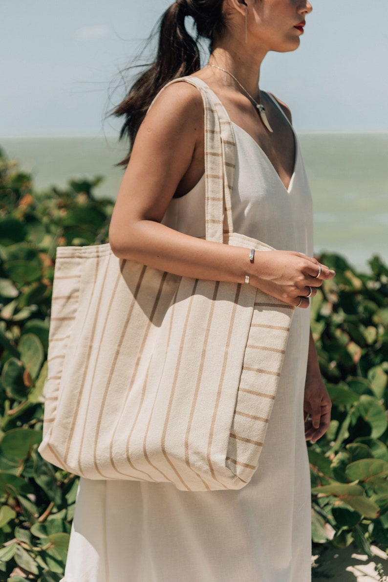 May include: A woman is carrying a large, striped tote bag with tan and white stripes. The bag is made of a natural fabric and has a relaxed, casual style.