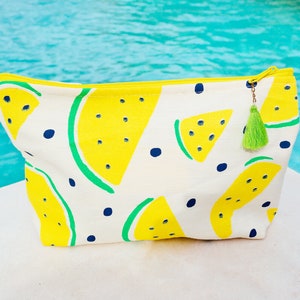 Puede incluir: Una bolsa de lona blanca con cremallera con estampado de sandía amarilla y un borla verde. La bolsa está sobre una superficie blanca.