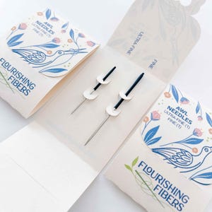 May include: Two fine, ultra-fine AWL needles with black handles and white accents are displayed in an open, cream-colored package. The package features a floral and bird design with the text "Flourishing Fibers" and "AWL NEEDLES".
