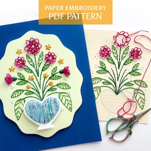 May include: A paper embroidery PDF pattern with a floral design. The design includes pink flowers, green leaves, and a blue heart-shaped vase. The text "PAPER EMBROIDERY PDF PATTERN" is at the top.