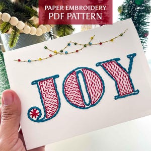 May include: A white card featuring the word JOY embroidered in red and white cross-stitch patterns with a teal outline. Above the text, a gold thread string displays small, colorful beads resembling holiday lights. The text PAPER EMBROIDERY PDF PATTERN is visible at the top.