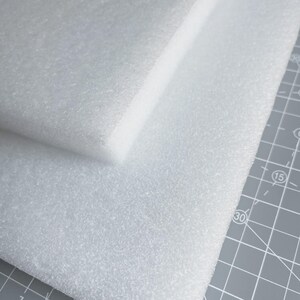 May include: Two white foam sheets, one stacked on top of the other. The foam has a textured surface and is likely used for packaging or crafting. The sheets are on a gray cutting mat.