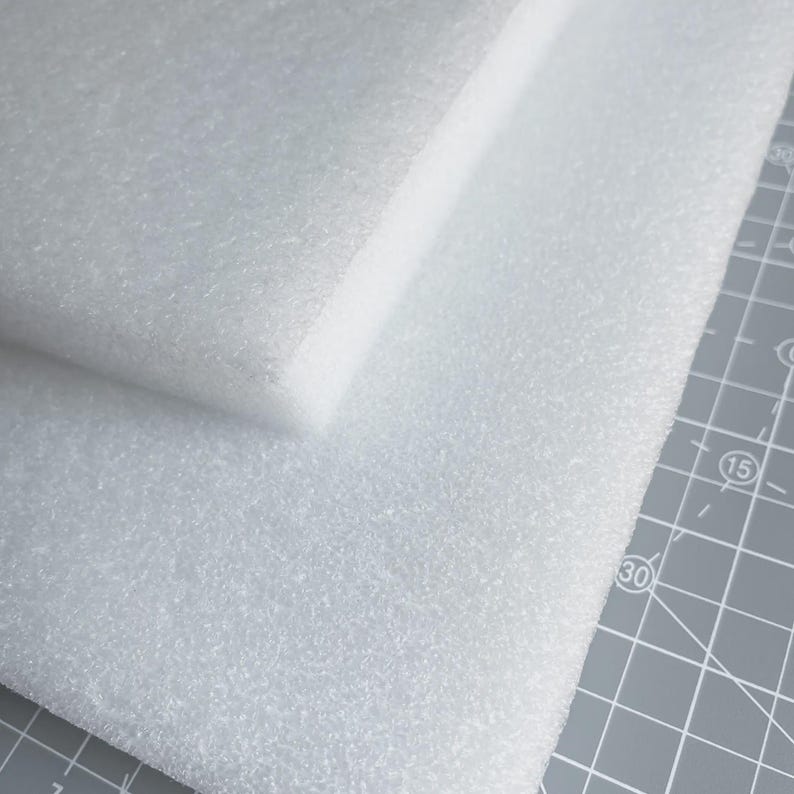 May include: Two white foam sheets, one stacked on top of the other. The foam has a textured surface and is likely used for packaging or crafting. The sheets are on a gray cutting mat.
