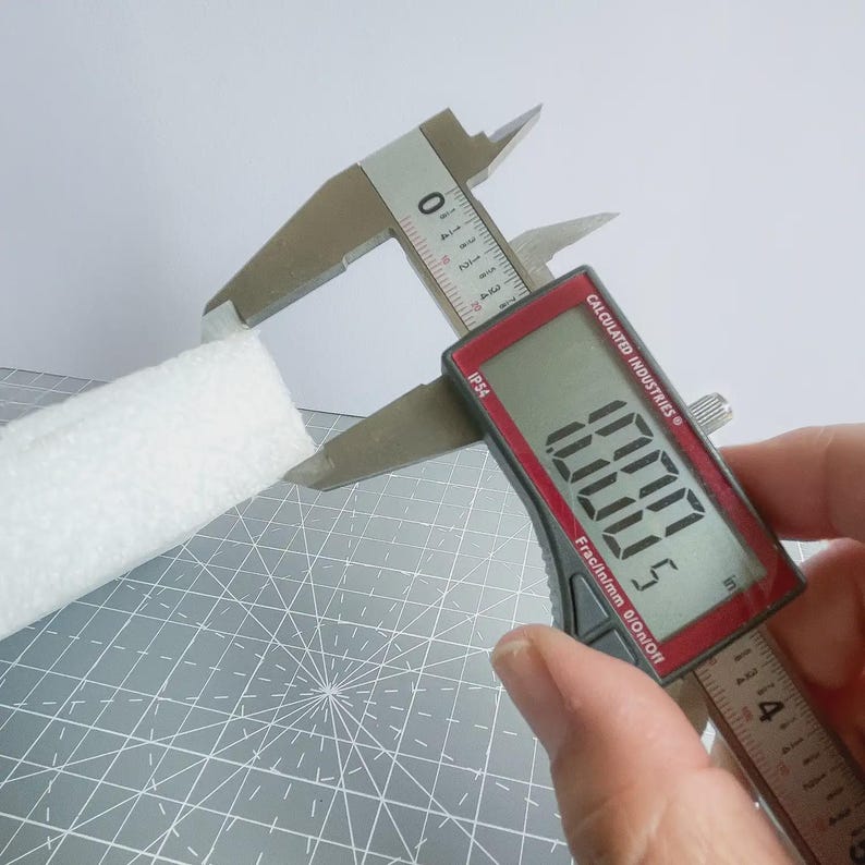 May include: A digital caliper measuring a white, textured rectangular object. The caliper's digital display reads 3.0005 inches. The caliper is silver and black with a red digital display. The background is a gray cutting mat.