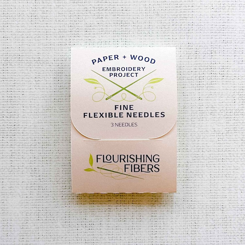 May include: A pink and white package of three fine flexible needles for embroidery. The package is labeled "Paper + Wood Embroidery Project" and "Flourishing Fibers".