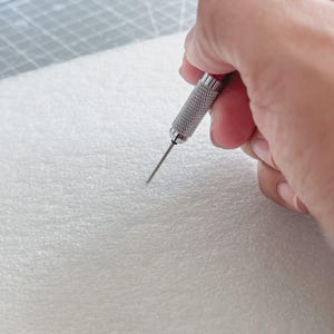 May include: A silver awl tool with a textured handle is held over a white, textured surface. The awl's sharp point is positioned to pierce the material. The background includes a gray cutting mat.