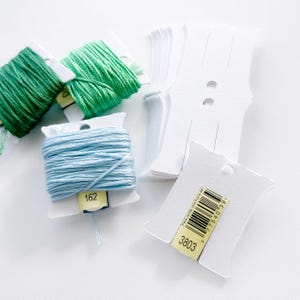 May include: A close-up of two spools of embroidery floss, one light blue and one green, with a white plastic label on each spool. The label on the blue spool has the number 162 printed on it. There are also several white plastic thread organizers with a yellow label that says "3803" and a barcode.