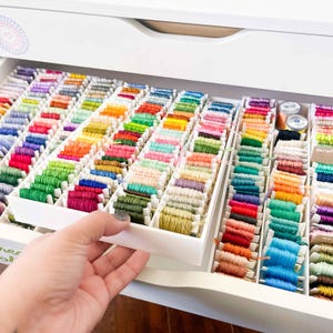 May include: A drawer filled with neatly arranged embroidery threads in a spectrum of colours, including pinks, greens, blues, and oranges. The threads are stored in white plastic trays, offering a tidy and visually pleasing setup for crafters.
