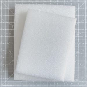 May include: Two white foam sheets, one slightly smaller, are stacked on a gray cutting mat. The foam has a textured surface. The cutting mat has a grid pattern with inch measurements.