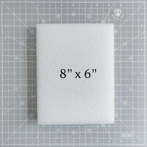 May include: A white, rectangular foam block, measuring 8 inches by 6 inches, is displayed on a gray cutting mat. The text "8" x 6"" is printed in black on the foam. The mat has grid lines and inch measurements.