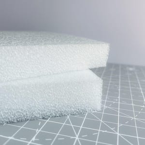 May include: Two white foam blocks with a textured surface are stacked on a gray cutting mat. The foam has a porous appearance, and the edges are clean and sharp. The background is a soft, neutral gray.