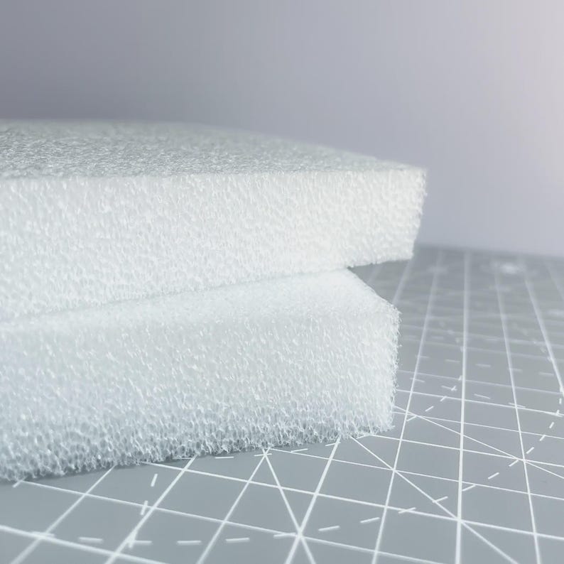 May include: Two white foam blocks with a textured surface are stacked on a gray cutting mat. The foam has a porous appearance, and the edges are clean and sharp. The background is a soft, neutral gray.