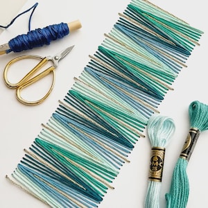 Puede incluir: Vista aérea de materiales de bordado. Un patrón diagonal de hilos azules, turquesas y dorados es el punto focal. Tijeras doradas, hilo azul en un carrete de madera y dos madejas de hilo de bordar DMC en turquesa y azul claro también son visibles.