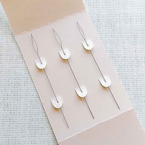 May include: A pack of six embroidery needles with white plastic holders. The needles are arranged in three rows of two, with the needles pointing upwards.