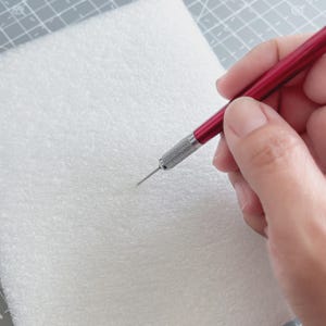 May include: A red and silver craft tool with a sharp needle tip is held over a white, textured surface. The tool has a metallic silver grip and a red handle. The image suggests a craft or hobby project.