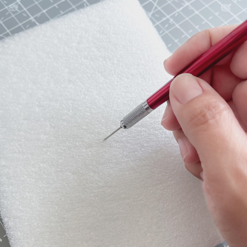 May include: A red and silver craft tool with a sharp needle tip is held over a white, textured surface. The tool has a metallic silver grip and a red handle. The image suggests a craft or hobby project.
