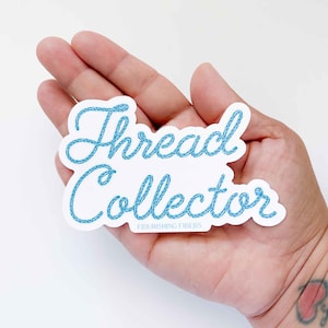 May include: A white sticker with blue thread lettering that reads "Thread Collector" with the words "Flourishing Fibers" below.