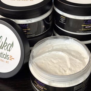 May include: Three jars of a white, creamy hair treatment product labeled "Alika Essentials - The Natural Me". The product is a Fenugreek Scalp Renewal Scrub that promotes a healthy environment for hair growth. The jars are black with a white label and purple text.