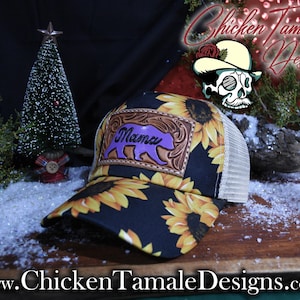 May include: A black trucker hat with a sunflower design and a beige mesh back. The front has a leather patch with the word "Mama" and a purple bear silhouette. The hat has a curved peak.