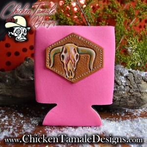 Könnte beinhalten: Ein pinkfarbener Neopren-Getränkekühler mit einem braunen Lederaufnäher, der mit einem bemalten Stierschädel verziert ist. Der Kühler trägt den Text "Chicken Tamale Designs" und die Website-Adresse www.ChickenTamaleDesigns.com.