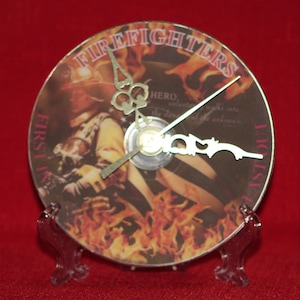 May include: A clock made from a CD with a firefighter image and the words "Firefighters First In Last Out" on it. The clock has gold hands and a clear plastic stand.