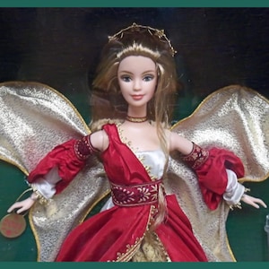 May include: A doll with blonde hair, wearing a red and white dress with gold accents. The doll has gold wings and a gold crown. A gold medallion hangs from the doll's wrist. The doll is in a green box.