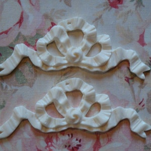 May include: Two white, decorative, plastic, ribbon-shaped embellishments with a ruffled design. The embellishments are perfect for adding a touch of elegance to crafts, home decor, or scrapbooking projects.