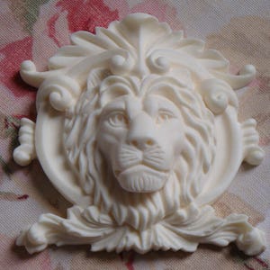 May include: White plaster lion head ornament with ornate scrollwork details. The lion's head is centered in a circular frame with a floral design.