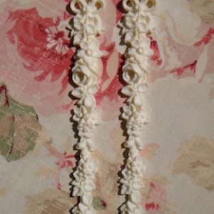 May include: Two white, decorative, floral-shaped trim pieces. Each piece is about 5 inches long and features a delicate, ornate design.