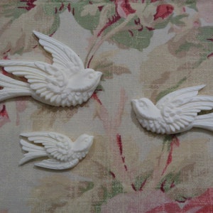 May include: Three white ceramic bird figurines with detailed wings and bodies. The birds are in flight and have a vintage style.