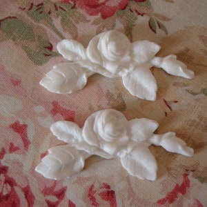 May include: Two white, rose-shaped embellishments with detailed petals and leaves. The floral accents are arranged on a floral patterned fabric background with pink and green hues. These decorative elements are ideal for craft projects.