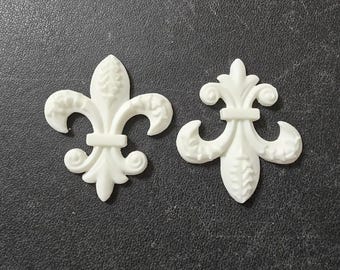 May include: Two white fleur-de-lis ornaments. The ornaments are made of a solid, opaque material and feature a detailed design with curved petals and a central vertical element. The background is a dark, textured surface.