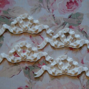 May include: Four white, decorative, plastic, ornate, ribbon-like embellishments with a ruffled design. The embellishments are arranged in a row on a floral fabric background.