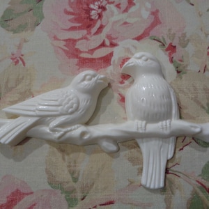 May include: Two white ceramic birds perched on a branch. The birds are facing each other and have detailed features. The branch is also made of ceramic and has a natural wood grain texture.