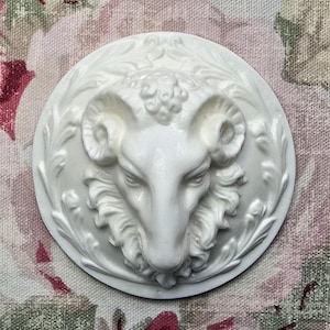 May include: White ceramic ornament featuring a detailed sculpted ram's head with horns and a floral design around the edges.