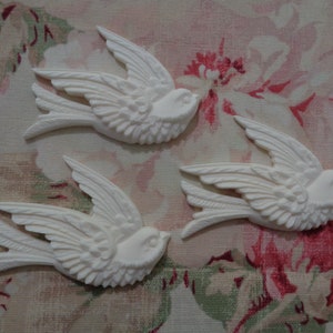 May include: Three white ceramic bird figurines with detailed wings and a textured surface. The birds are arranged in a staggered pattern on a floral fabric background.