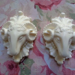 May include: Two white ceramic ram head ornaments with detailed features, including horns, eyes, and a textured surface. The ornaments are set against a floral background.